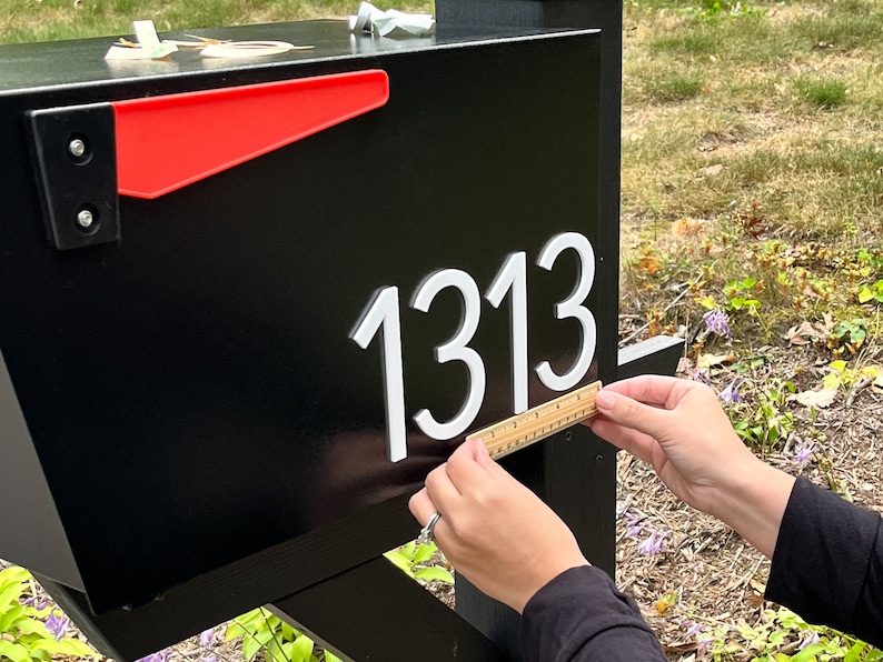 May include: A black mailbox featuring a bright red flag and the white house numbers 1313 attached to the side. A small wooden ruler is held against the bottom of the numbers to show their scale, measuring approximately 3 inches in height.