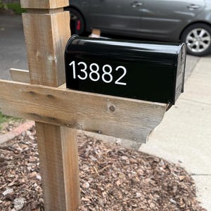 May include: A black metal mailbox with the white numbers 13882 displayed on the side. The mailbox is mounted on a weathered, light brown wooden post structure that features a vertical support beam and a horizontal shelf for the mailbox to rest on.