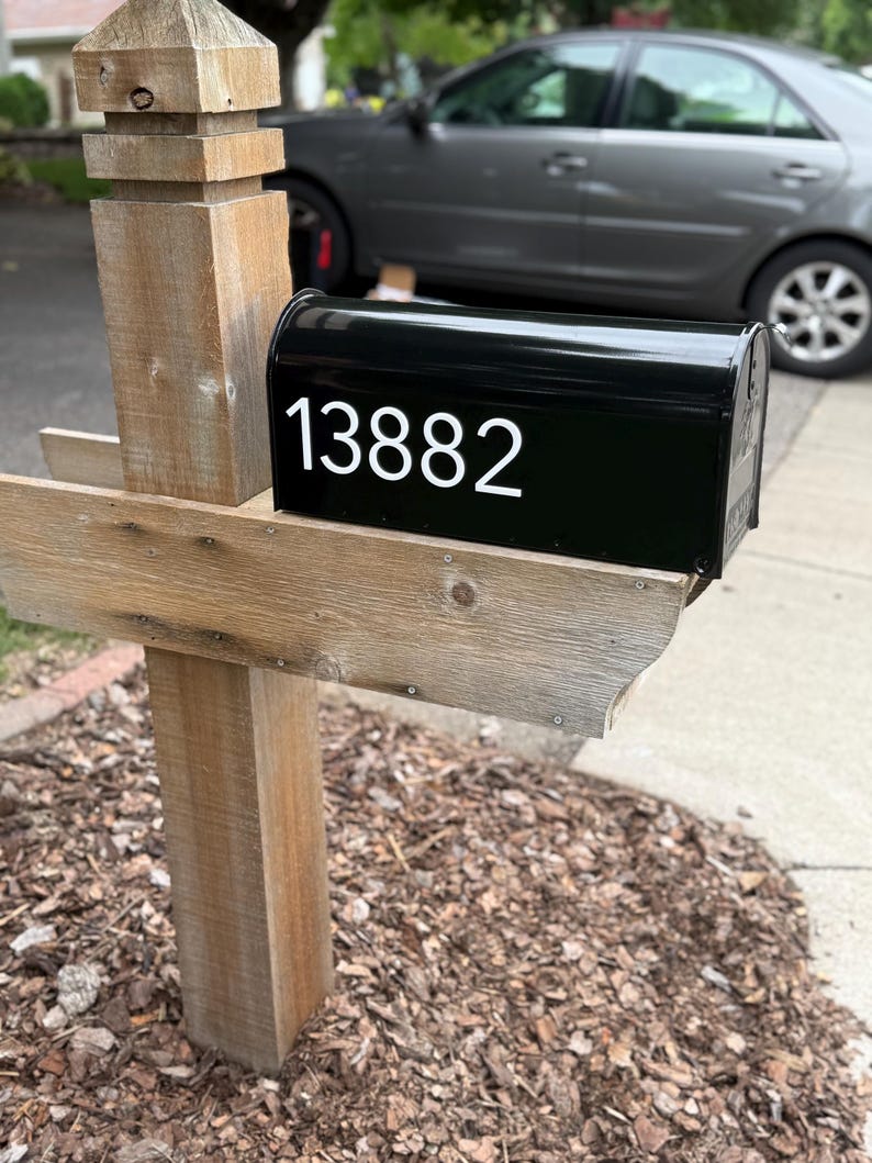 May include: A black metal mailbox with the white numbers 13882 displayed on the side. The mailbox is mounted on a weathered, light brown wooden post structure that features a vertical support beam and a horizontal shelf for the mailbox to rest on.