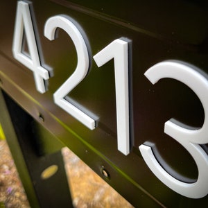 May include: A dark brown metal surface featuring the white house numbers 4213 in a clean, sans-serif font. The numbers are raised and have a distinct depth, creating a clear contrast against the dark background of the mailbox or sign structure.