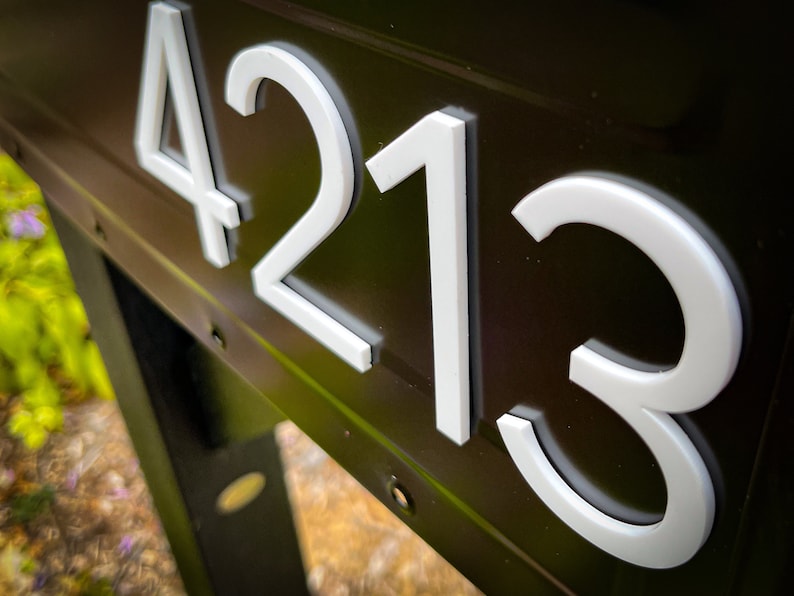 May include: A dark brown metal surface featuring the white house numbers 4213 in a clean, sans-serif font. The numbers are raised and have a distinct depth, creating a clear contrast against the dark background of the mailbox or sign structure.