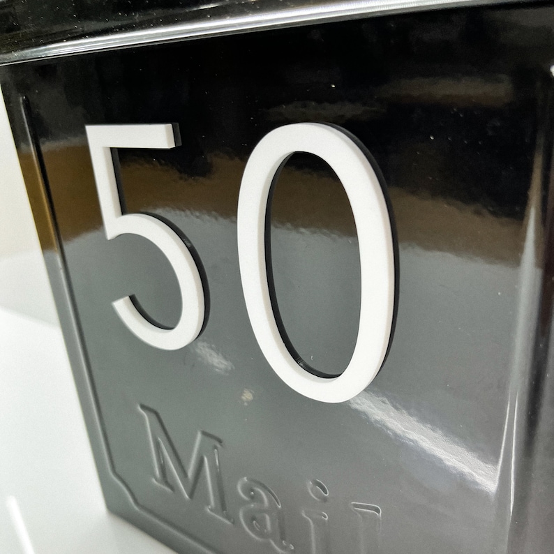 May include: A glossy black mailbox featuring the white number 50 prominently displayed on the front. Below the number, the word Mail is embossed into the surface of the metal box, which has a reflective finish and clean, modern lines.