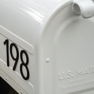 May include: A glossy white metal mailbox featuring the black house numbers 198 on the side. The front door panel is embossed with the text U.S. MAIL Approved By The Postmaster General and includes a white handle at the top for opening the lid.