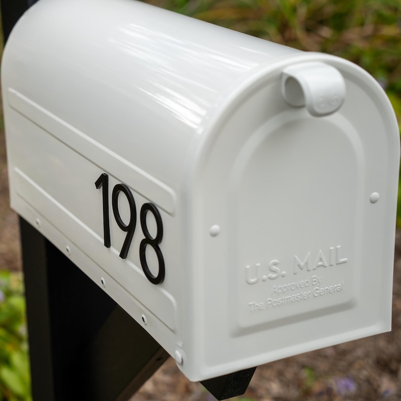 May include: A white metal mailbox featuring the black house number 198 on the side. The front door is embossed with the text U.S. MAIL and Approved By The Postmaster General. The mailbox is mounted on a black post and has a curved top design.