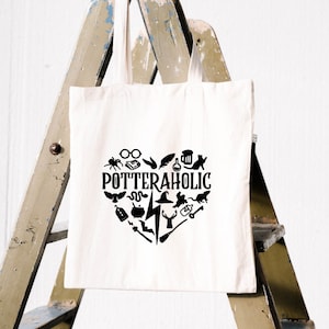 May include: A cream-coloured tote bag featuring a black heart-shaped design. The heart is filled with icons and the word "POTTERAHOLIC". The bag hangs on a wooden ladder against a white wall.