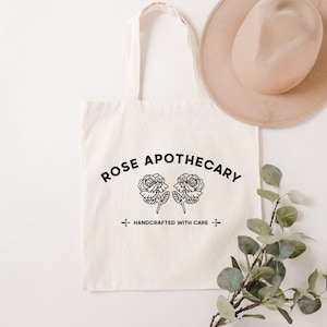 May include: A white canvas tote bag with black text that reads "ROSE APOTHECARY" in an arc above two stylized roses. The text "HANDCRAFTED WITH CARE" is centered below the roses.