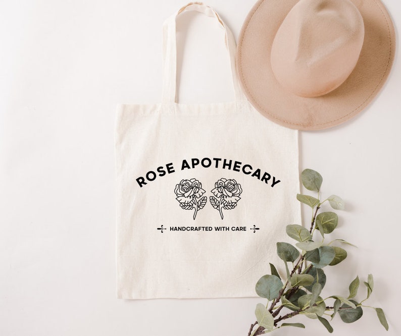 May include: A white canvas tote bag with black text that reads "ROSE APOTHECARY" in an arc above two stylized roses. The text "HANDCRAFTED WITH CARE" is centered below the roses.