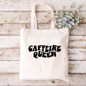 May include: A white canvas tote bag with black text that reads "Caffeine Queen" in a retro font.