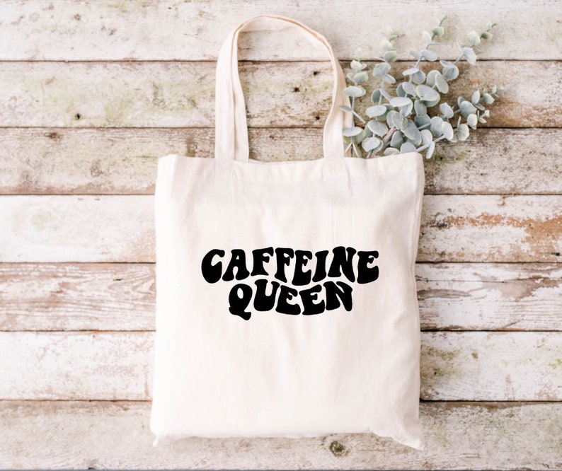 May include: A white canvas tote bag with black text that reads "Caffeine Queen" in a retro font.