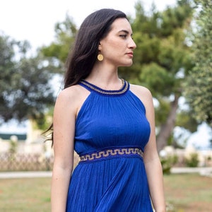 May include: A woman wearing a royal blue maxi dress with gold trim and a gold circle earring. The dress has a halter neckline and a pleated skirt.