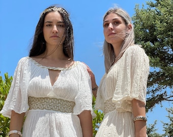 Peut inclure: Deux femmes portant des robes blanches et fluides avec des broderies dorées. Les robes ont un décolleté en V et des manches courtes. Les femmes sont debout devant un ciel bleu et des arbres verts.