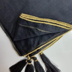 May include: A black scarf with gold trim and tassels. The scarf is made of a soft, flowing fabric and features a decorative gold border along the edges. Black tassels hang from the corners, adding a touch of elegance.