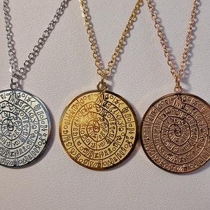 Puede incluir: Tres collares con colgantes circulares que presentan un diseño en espiral y letras intrincadas. Los colgantes son de plata, oro y oro rosa.