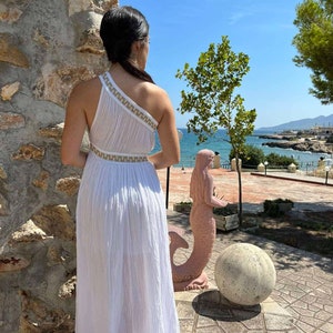 Pode incluir: Um vestido branco maxi com detalhes dourados, uma alça única e uma saia fluida. O vestido é usado por uma pessoa em pé em frente a uma parede de pedra e olhando para o oceano.
