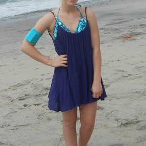 Pu&ograve; includere: Una giovane persona indossa un vestito estivo blu con un top bikini a stampa floreale sotto. &Egrave; in piedi su una spiaggia sabbiosa con l'oceano sullo sfondo.