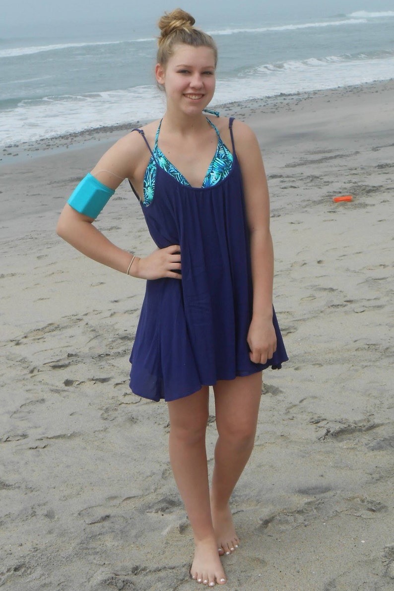 Pu&ograve; includere: Una giovane persona indossa un vestito estivo blu con un top bikini a stampa floreale sotto. &Egrave; in piedi su una spiaggia sabbiosa con l'oceano sullo sfondo.