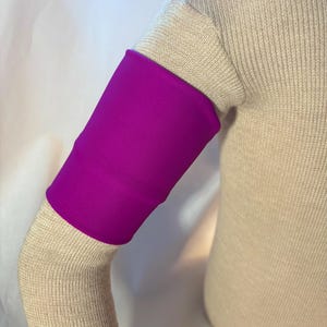 May include: A vibrant magenta compression sleeve worn on an arm. The sleeve is a solid color and appears to be made of a smooth, stretchy material. The arm is covered in a light beige knitted fabric.