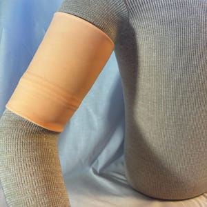 May include: A close-up of a peach-colored arm sleeve worn over a gray sweater. The sleeve is made of a soft, stretchy material and is designed to cover the entire arm from the wrist to the elbow.