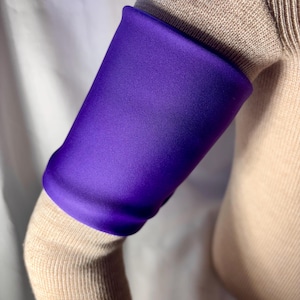 May include: A close-up of a purple arm warmer made of a soft, stretchy fabric. The arm warmer is worn on the left arm of a person wearing a beige sweater.