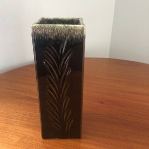 May include: A square ceramic vase with a dark green glaze and a light green rim. The vase has an embossed design of a leafy branch on the front.