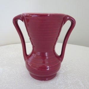 May include: A maroon-colored vase with two curved handles. The vase has a wide, rounded base that tapers inward before flaring out again towards the top. The handles curve gracefully from the top of the vase, creating an elegant silhouette. The vase has a glossy finish.