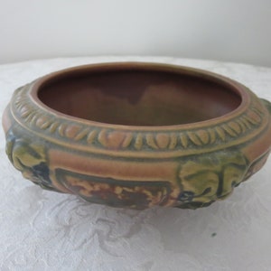May include: A decorative, antique ceramic bowl with a wide rim and a detailed, embossed design. The bowl features a color palette of brown, green, and tan. The rim has a repeating pattern, and the sides have sculpted floral elements.