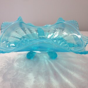 May include: A light blue, vintage glass bowl with a scalloped rim and a detailed, ribbed design. The bowl is supported by three small, rounded feet. This decorative piece is ideal for serving or display.