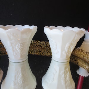May include: Two white milk glass candle holders with a grapevine design. The holders are sitting on a gold metal tray with a decorative design.