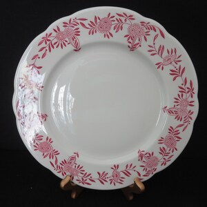 May include: A white ceramic dinner plate with a pink floral design around the rim. The plate has a scalloped edge.