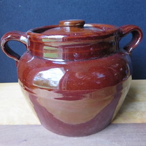May include: A brown glazed ceramic bean pot with two handles and a lid. The pot has a rounded body and a slightly flared rim.