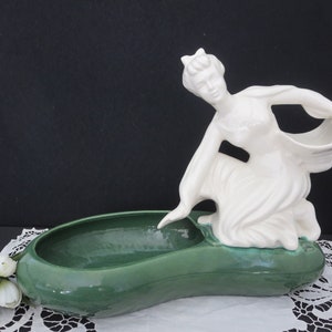 May include: A white ceramic figurine of a woman holding a basket, attached to a green ceramic planter. The planter is shaped like a long, oval bowl.