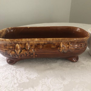 May include: A brown ceramic planter with a decorative floral design and four feet. The planter is oval shaped and has a glazed finish.