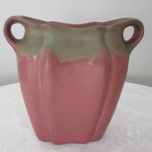 May include: A pink ceramic vase with a square base and two loop handles. The top edge and handles are a muted green. The vase has a textured surface and a slightly flared opening. A decorative piece for the home.