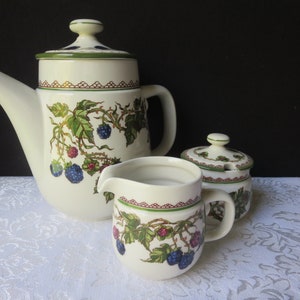 May include: A white teapot, creamer, and sugar bowl with a green and blue blackberry pattern. The teapot has a lid and a handle. The creamer and sugar bowl have handles.