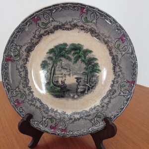 May include: A vintage ceramic plate with a black and white landscape scene featuring a building, trees, and a river. The plate has a decorative border with a repeating pattern of green leaves and red flowers.
