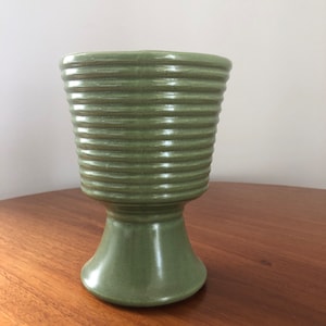 May include: A green ceramic planter with a ribbed design and a pedestal base.