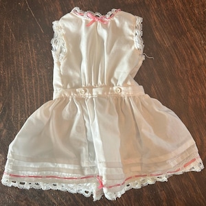 May include: A vintage white doll dress with a pleated skirt and a fitted bodice. The dress features delicate lace trim along the neckline and hem, with pink ribbon accents. Two small buttons adorn the waistline, and a pink bow is at the neckline.