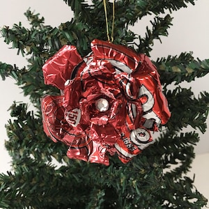 May include: A handmade Christmas ornament shaped like a flower, crafted from recycled red aluminum cans. The ornament has a silver center and is hanging from a gold string. The ornament is displayed on a green Christmas tree.
