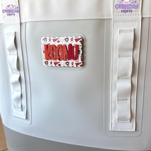 May include: A light gray tote bag featuring white vertical webbing straps and a rectangular patch with a scalloped edge. The patch displays a red baseball-themed pattern and the name KEANU in bold red letters. A small logo reads CAROLCOW CRAFTS.