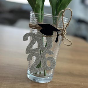 May include: A clear glass vase with green stems and a silver glitter "2026" decoration and a black graduation cap. The vase is wrapped with twine. The background is a light brown wooden surface.