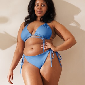 Puede incluir: Un conjunto de bikini azul claro. La parte superior es de estilo triangular con lazos ajustables, y la parte inferior es de corte clásico con lazos laterales. El traje de baño está hecho de una tela lisa de color liso. La modelo está de pie frente a un fondo neutro.