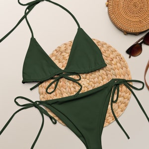 Peut inclure: Un ensemble de bikini vert foncé avec un haut triangle et un bas taille haute. Le bikini est présenté sur un tapis de paille tissé avec un sac rond en paille et des lunettes de soleil.