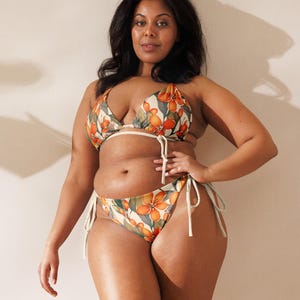 Könnte beinhalten: Eine Frau trägt ein Bikini-Oberteil und -Unterteil mit floralem Muster und weißen Bändern. Der Bikini ist ein Dreiecks-Oberteil mit einem Halfter-Band und einem tief sitzenden Unterteil.