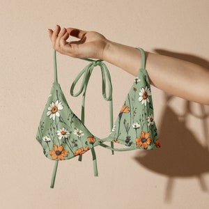 May include: A green bikini top with a floral print featuring white and orange flowers. The top has adjustable straps and a tie closure at the back.