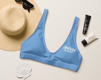 May include: A light blue bikini top with the word "CANCUN" and the date "6.5.2025" printed in white. The top is next to a straw hat, sunglasses, sunscreen, and a seashell.
