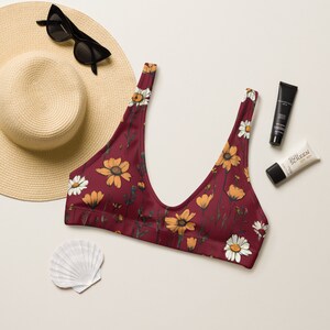 Puede incluir: Un top de bikini rojo con estampado floral con flores amarillas y blancas. El top tiene un escote redondo y tirantes ajustables.