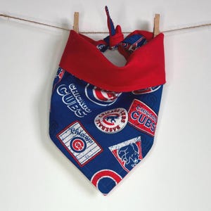 May include: A blue and red dog bandana with the Chicago Cubs logo and other baseball-themed graphics. The bandana is tied with a red bow.