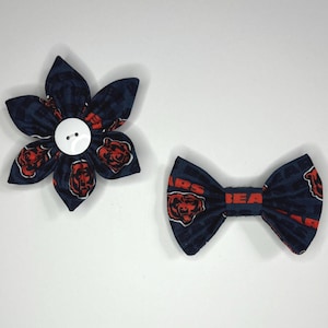 May include: A navy blue bow tie and flower hair clip with a Chicago Bears logo print. The bow tie and flower clip are both made of fabric and have a white button.