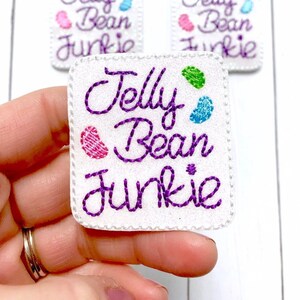 May include: White felt patch with purple embroidery that reads "Jelly Bean Junkie". The patch has three colorful jelly bean shapes embroidered on it.