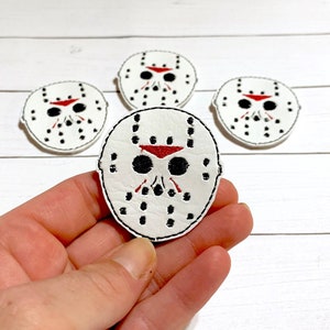 May include: Four white embroidered iron-on patches of a hockey mask with red and black details. The patches are on a white wooden surface.